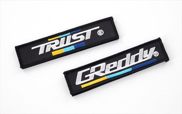 Close-up of detachable GREDDY and TRUST logo patches included with the GREDDY Backpack 45L, showcasing brand customization options.